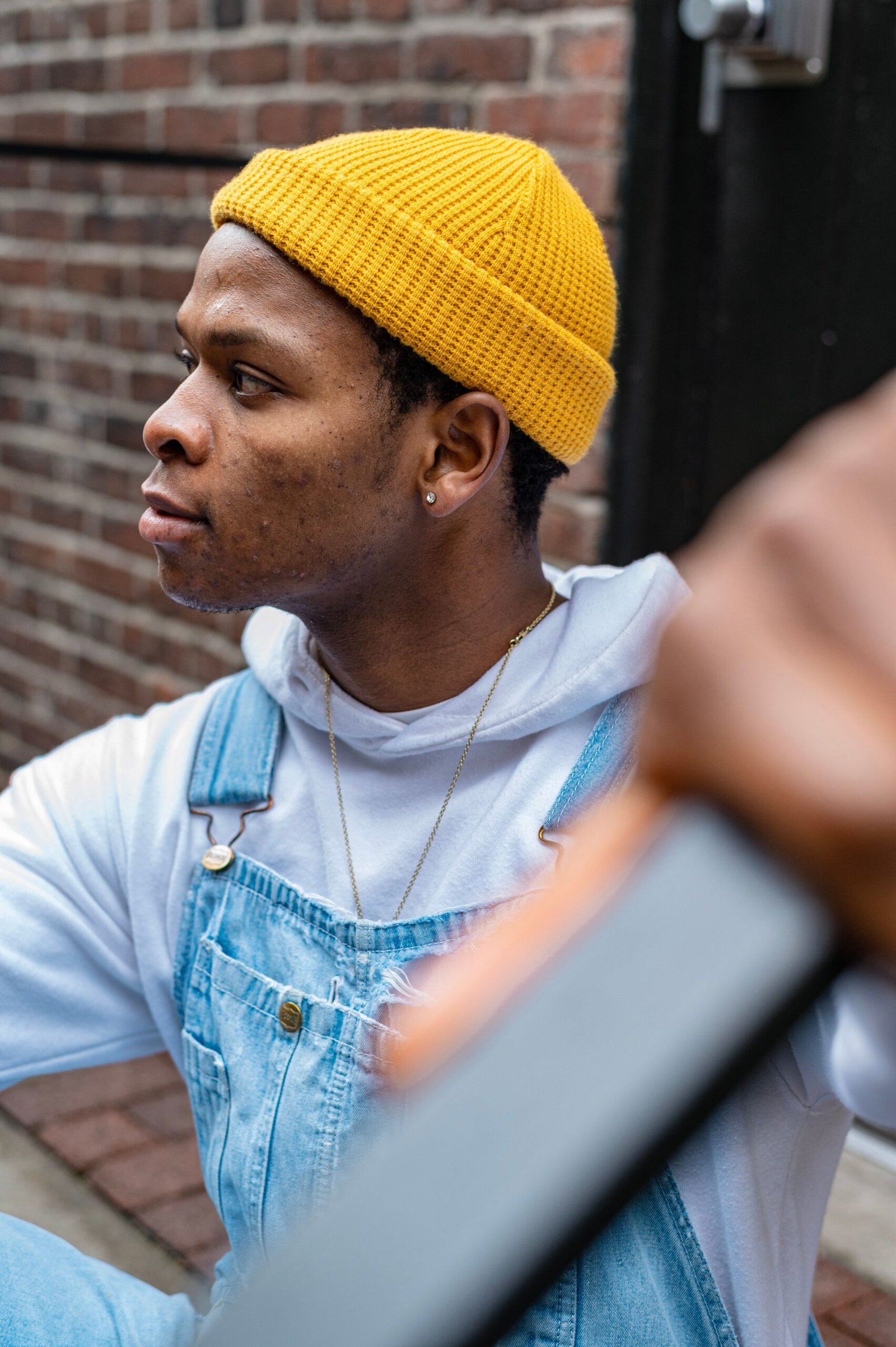 Stylish young man wearing denim overalls and a yellow beanie in an urban setting.